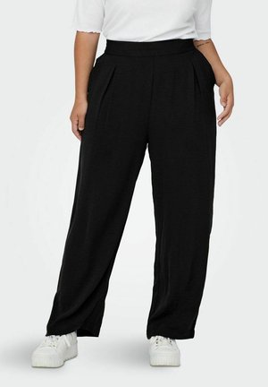 Femme portant un pantalon large noir avec des poches, un haut blanc côtelé et des baskets blanches à plateforme, debout devant un fond blanc.