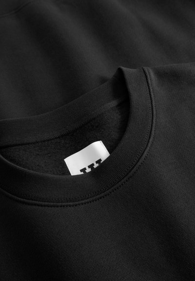 Sweat-shirt noir à col rond, texture lisse et détails de coutures visibles. L'étiquette présente un fond blanc avec un texte noir.