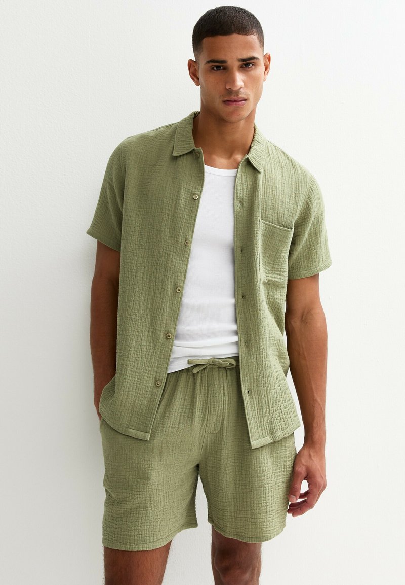 Camicia a maniche corte di colore verde oliva chiaro, con tessuto texturizzato, chiusura a bottoni e tasca sul petto, abbinata a pantaloni corti coordinati con vita elastica.