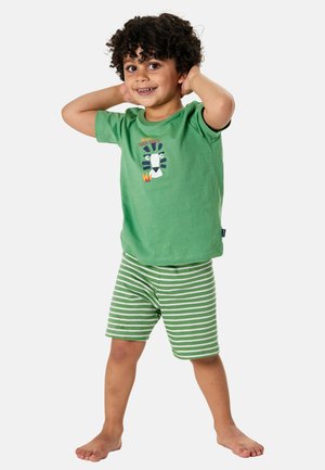 Smiling child with curly hair wearing green “Cool Tiger Vibes” t-shirt and green-and-white striped shorts, standing barefoot with hands behind head.