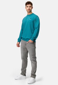 Teal sweatshirt med ribbede manchetter, grå jeans og hvide sneakers. Sweatshirten har et logo på venstre brystområde. Casual pasform.
