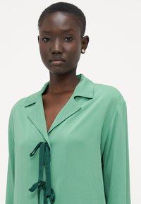 Lichtgroene blouse met een kraag en striksluiting aan de voorkant. Glad materiaal en een losse pasvorm, met een lichte glans.