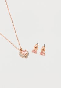 Σετ κολιέ και σκουλαρίκια καρφάκια σε ροζ χρυσό με ροζ πέτρες σε σχήμα καρδιάς και μικρά διαφανή κρυστάλλινα στοιχεία σε αλυσίδα και καρφίτσες, πάνω σε λευκό φόντο.