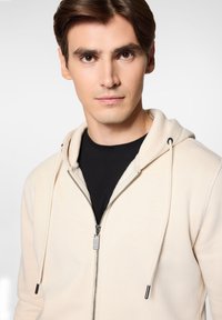 Kremowa bluza z kapturem, z zamkiem błyskawicznym z przodu, sznurkiem do regulacji oraz czarnymi akcentami. Noszona na czarnej koszulce, z delikatnym logo na piersi.