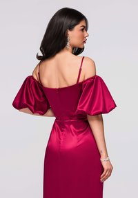 Robe longue en satin bordeaux avec manches bouffantes décolletées, corsage ajusté et fermeture éclair au dos. Accent de bracelet argenté visible.