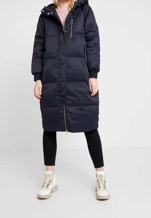 Femme portant un long manteau matelassé noir avec fermeture éclair à l'avant, des leggings noirs et des bottines blanches à lacets, debout devant un fond uni.