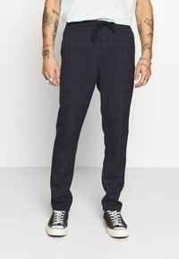 Marineblauwe geruite broek met rode lijnen, gemaakt van zachte stof, met een elastische tailleband en een trekkoord, gecombineerd met zwarte sneakers.