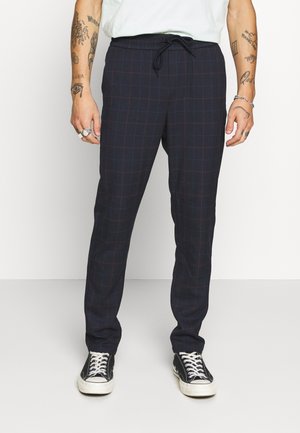 Pantalons à carreaux navy avec des lignes rouges, en tissu doux, dotés d'une taille élastique et d'un cordon de serrage, associés à des baskets noires.