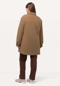 Brun fleecejacka med krage, lös passform och knäppte ärmslut, kombinerad med bruna byxor och beige skor med gröna detaljer.