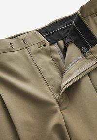 Pantaloni khaki con chiusura a zip e gancio, dotati di una vita nera e dettagli argento. Tessuto liscio con pieghe nella parte anteriore.