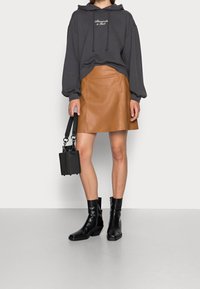 Donna che indossa un hoodie grigio scuro, una gonna di pelle beige, stivaletti neri alla caviglia, e porta una piccola borsa nera su uno sfondo semplice.
