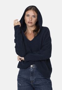 Maglione con cappuccio blu navy a V, realizzato in morbido tessuto testurizzato, con maniche lunghe e un taglio casual. Abbinato a jeans strappati.