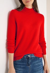 Roter Strickpullover mit hohem, geripptem Kragen und umgeschlagenen Ärmeln, kombiniert mit blauer Jeans. Der Pullover hat eine lockere Passform und einen gerippten Saum.