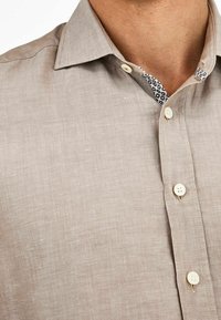 Camicia in lino marrone chiaro con colletto classico, piccoli bottoni bianchi e tessuto a fantasia a contrasto all'interno del colletto.