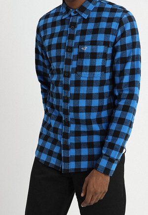 Man draagt een blauw-zwart geruit overhemd met lange mouwen, een borstzak en zwarte broek.