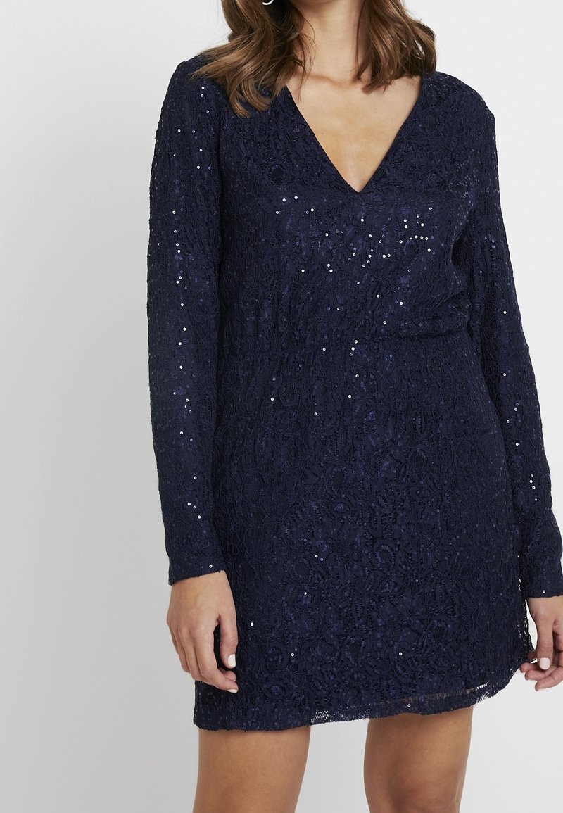 Robe en dentelle marine à manches longues et décolleté en V profond, ornée de sequins pour plus de texture et de détails. Longueur au-dessus du genou.