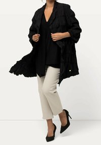 Woman wearing a textured black jacket and top, beige ankle-length pants, and black high-heeled shoes, standing with one leg slightly raised.