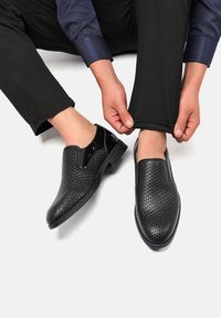 Scarpe nere slip-on con texture a motivo intrecciato e dettagli lucidi. Presentano una punta arrotondata e una suola piatta, abbinate a pantaloni neri sartoriali.