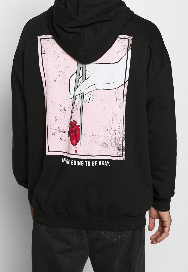 Una sudadera negra muestra un gráfico de una mano blanca sosteniendo un corazón rojo que gotea con palillos sobre un fondo rosa, el texto dice "Vas a estar bien."