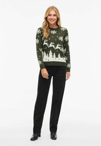 Woman wearing a dark green Christmas sweater with white reindeer, snowflakes, and trees, paired with black pants and black boots.