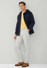 Giacca blu navy con cappuccio e due tasche frontali, camicia gialla sotto, pantaloni grigio chiaro e scarpe in suede marroni. Sfondo neutro.