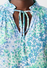 Blouse à fleurs dans des tons bleus et verts, avec un décolleté en V et un détail noué. Elle présente un col froncé et un tissu doux et léger avec un motif texturé.