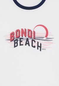 Weißes Baumwoll-T-Shirt mit runder marinefarbener Kragen, auf dem "BOND" in Rot, "BEACH" in Schwarz und eine rote Sonnengrafik über welligen pinken Linien abgebildet sind.