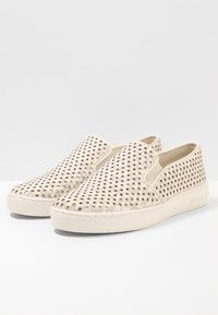 Chaussures slip-on avec tige en toile beige décorée de découpes en forme d'étoiles dorées, finition texturée et semelle en caoutchouc blanche. Design simple et léger.