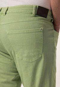 Pantalones de mezclilla verde con un diseño ajustado, bolsillos traseros distintivos y un detalle de parche de cuero marrón en la cinturilla.