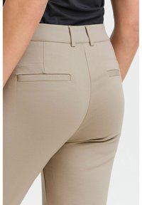 Béžové nohavice na mieru s hladkým povrchom, vyznačujúce sa dekoratívnymi zadnými vreckami a priliehavým strihom. Navrhnuté pre elegantný vzhľad.