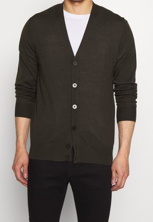 Mann trägt einen dunkelbraunen, geknöpften Cardigan über einem weißen Hemd und schwarzen Hosen, steht mit entspannt an den Seiten hängenden Armen.