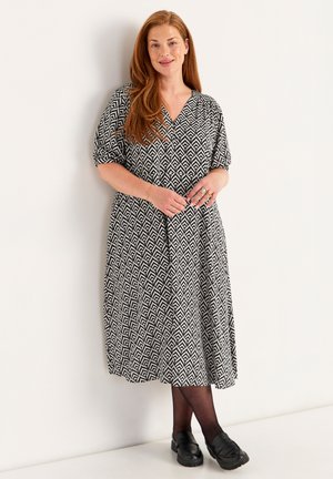 Schwarz-weiß gemustertes Kleid mit kurzen Ärmeln, V-Ausschnitt und fließendem Schnitt, kombiniert mit schwarzen Loafers und schwarzen Strumpfhosen.