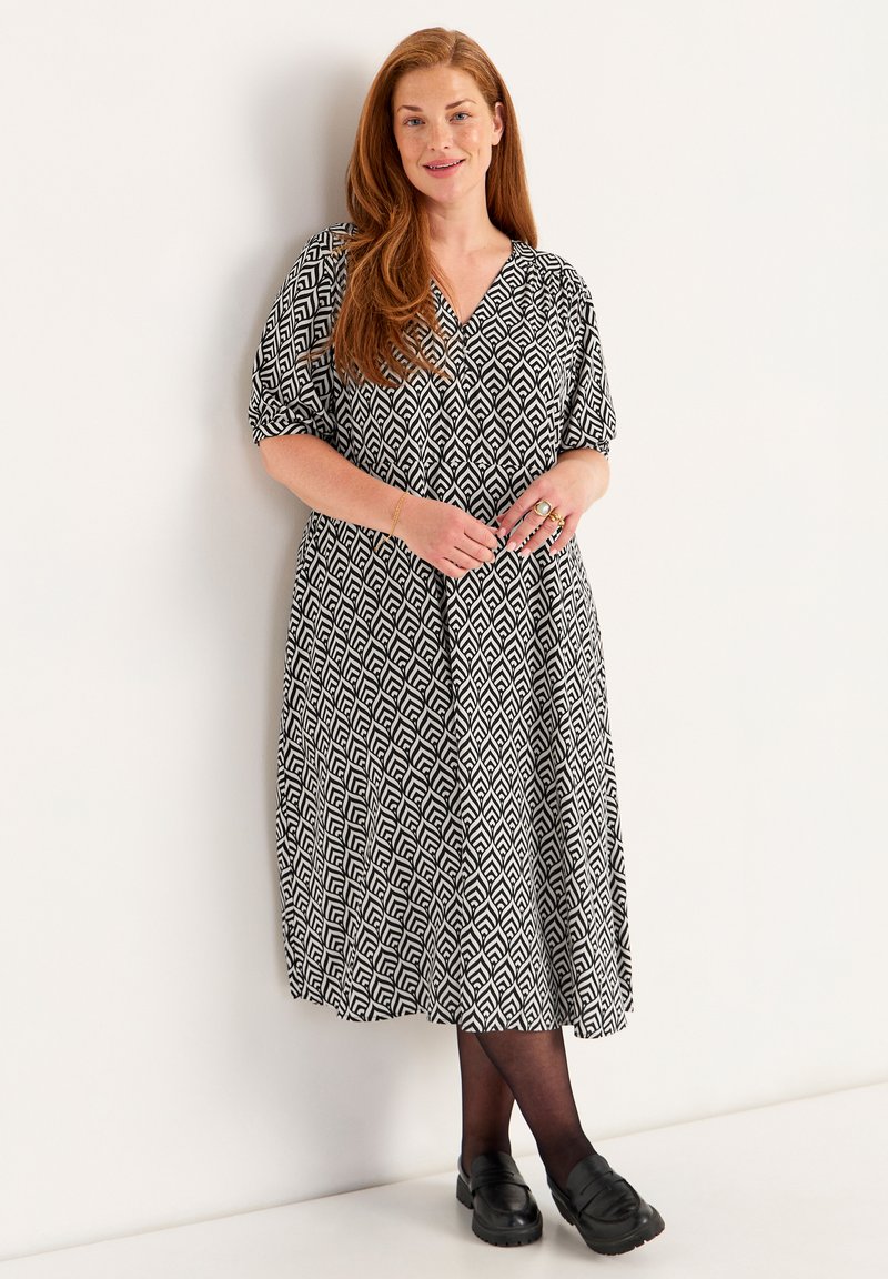 Mekko, jossa on mustavalkoinen kuvio, lyhyet hihat, V-kaula-aukko ja virtaava muotoilu, yhdistettynä mustiin loafer-kenkiin ja mustiin sukkahousuihin.