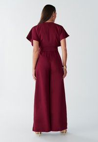 Mono color burdeos con mangas cortas acampanadas y cintura con cinturón; piernas anchas y textura suave; detalle sutil de cremallera trasera.