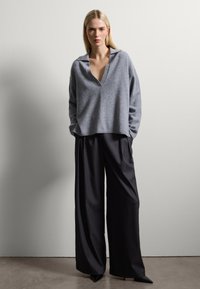 Grauer Strickpullover mit V-Ausschnitt und Kragen, kombiniert mit hoch geschnittenen, weit geschnittenen schwarzen Hosen. Sanfte Texturen, schnörkelloses Design.