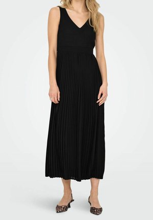 Femme portant une robe midi plissée noire sans manches avec un col en V et des chaussures à talons bas imprimé léopard, debout sur fond blanc.