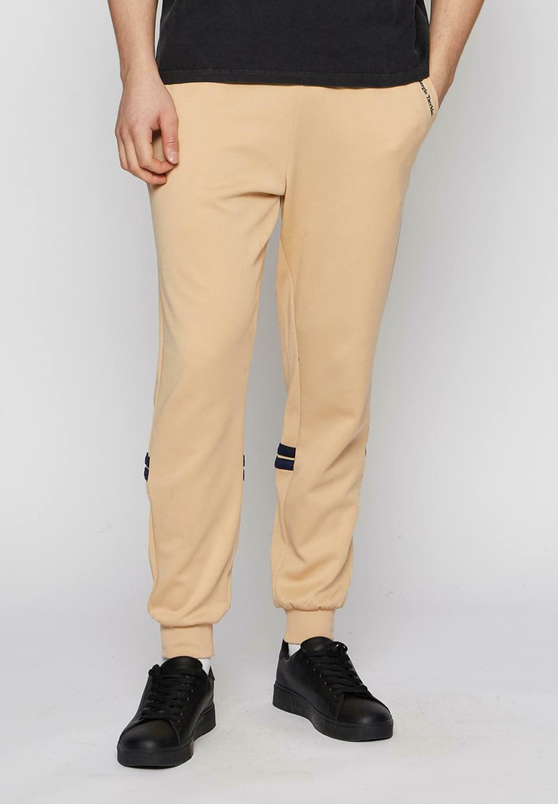 Beige sweatpants med en tapered design, elastiske manchetter og marineblå striber nær kanten. Båret med sorte sneakers.