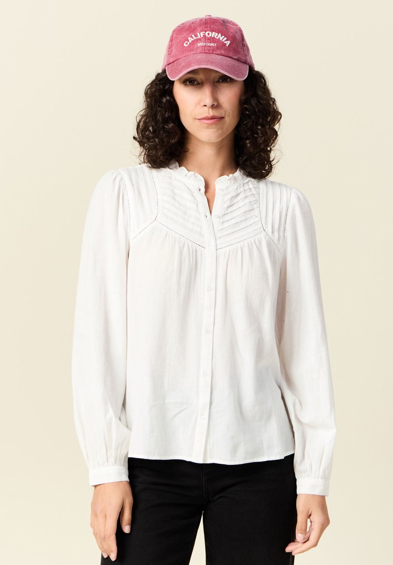 Weiße Bluse mit langen Ärmeln und Knopfleiste, mit strukturierten Falten an den Schultern und einem rüschenden Kragen, kombiniert mit einer rosa Mütze mit der Aufschrift "CALIFORNIA."
