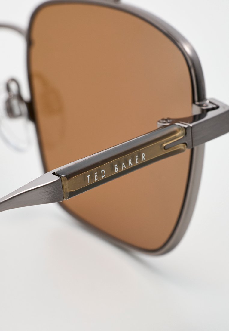 Braun getönte Sonnenbrille mit einem Metallrahmen, ausgestattet mit einer dünnen Augenbrauenstange und Akzenten in einem Grauton. "Ted Baker"-Logo am Bügel.