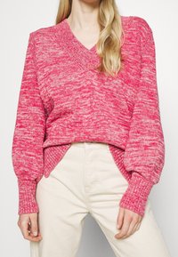 Roze gebreide v-halstrui met een gemarmerde textuur, ballonnenmouwen en zijsplitten. Gecombineerd met lichte broek.