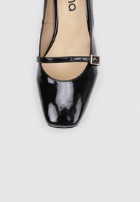 Zapato de charol negro con punta cuadrada, que cuenta con una correa fina y un acento de hebilla dorada. Textura suave con un acabado brillante.