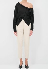 Blusa nera off-shoulder con chiusura a bottoni e orlo diagonale, abbinata a pantaloni beige aderenti e tacchi neri a punta. Tessuto liscio.