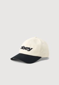 Béžová bavlnená čiapka s čiernym okrajom. Obsahuje logo "obey" v tučnom čiernom písmene. Zahrňuje odvetrávacie dierky a zaoblený dizajn koruny.