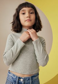 Grå ribbet langærmet top med højt halsudskæring, parret med højhårede lyseblå denimshorts. Baggrunden har en gul cirkulær form.