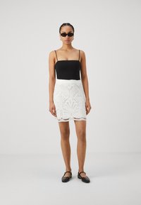 Haut noir ajusté avec des bretelles spaghetti, associé à une jupe en dentelle blanche avec des motifs floraux et un ourlet festonné. Chaussures plates noires.
