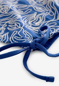 Modrý textil s bílou texturovanou výšivkou zobrazující tekoucí vzor. V rohu je modrá šňůrka na zavázání pro uzavření.