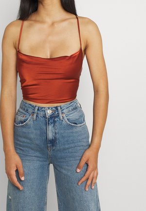 Femme portant un crop top en satin couleur rouille à fines bretelles associé à un jean large taille haute bleu clair, debout devant un fond uni.