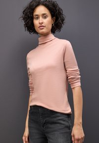 Helles rosa Oberteil mit hohem Kragen aus weichem Stoff und langen Ärmeln, kombiniert mit dunkelgrauen Jeans. Strukturiert Oberfläche auf dem Oberteil sichtbar.