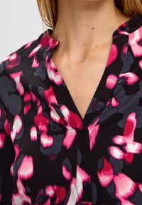 Primer plano de una blusa negra con cuello en V que presenta patrones florales abstractos en tonos vibrantes de rosa, blanco y gris en la parte superior del pecho y el cuello de una persona.