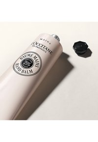 Roze tube van L'Occitane handbalsem met zwarte accenten, een zilverkleurige dop en een ronde sticker met de tekst "25% sheaboter." De dop ligt naast de tube op een beige achtergrond.
